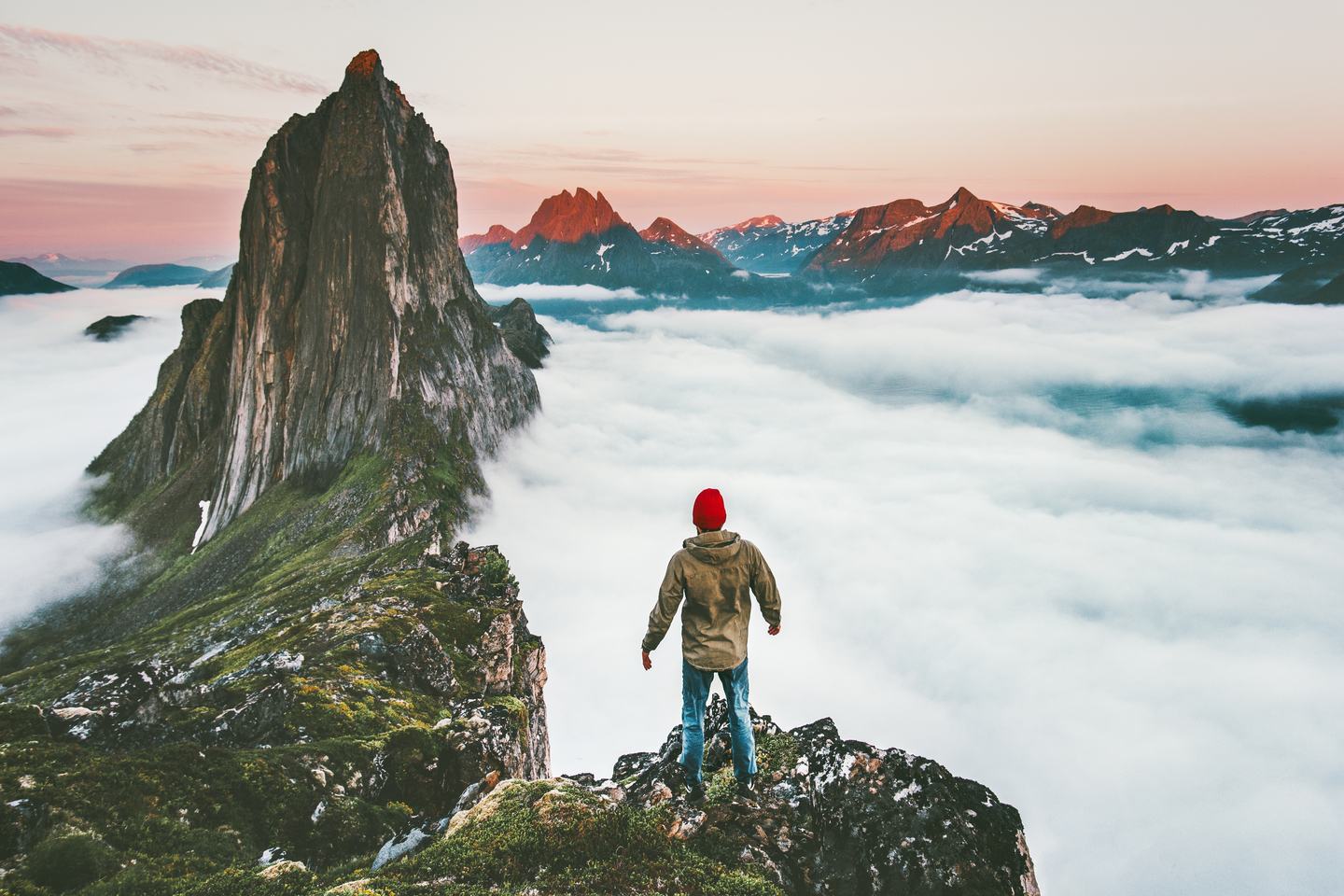A figure standing on a ridge above a sea of cloud, facing Segla and the Norwegian peaks at sunrise