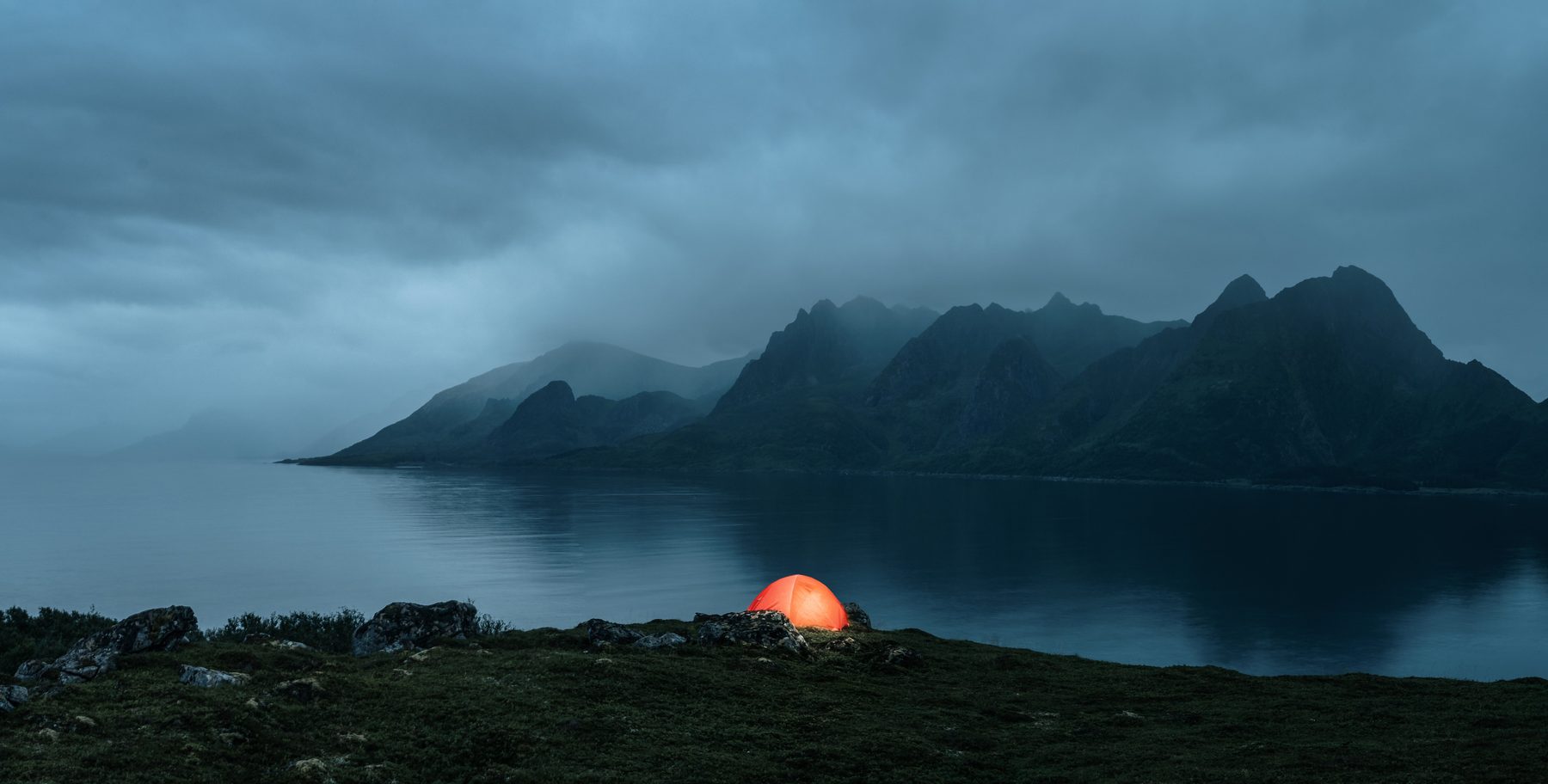 A lit orange tent on a headland above a fjord at dusk, with a mountain ridge under low cloud beyond