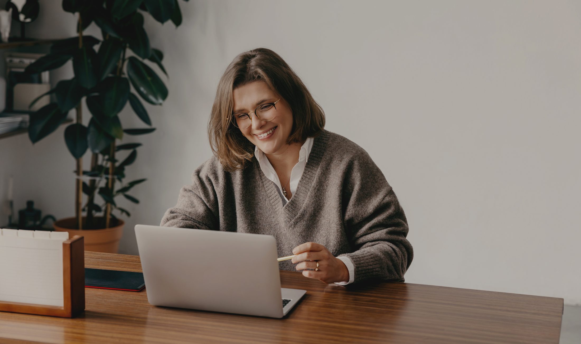 A woman in glasses smiling at her laptop at a wooden desk