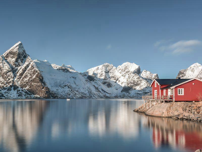 A red fishing cabin at the water's edge in Lofoten, snow-covered peaks mirrored in the still fjord