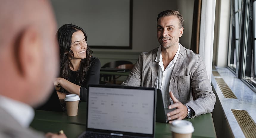 Two colleagues talking across a meeting table with a dashboard open on a laptop